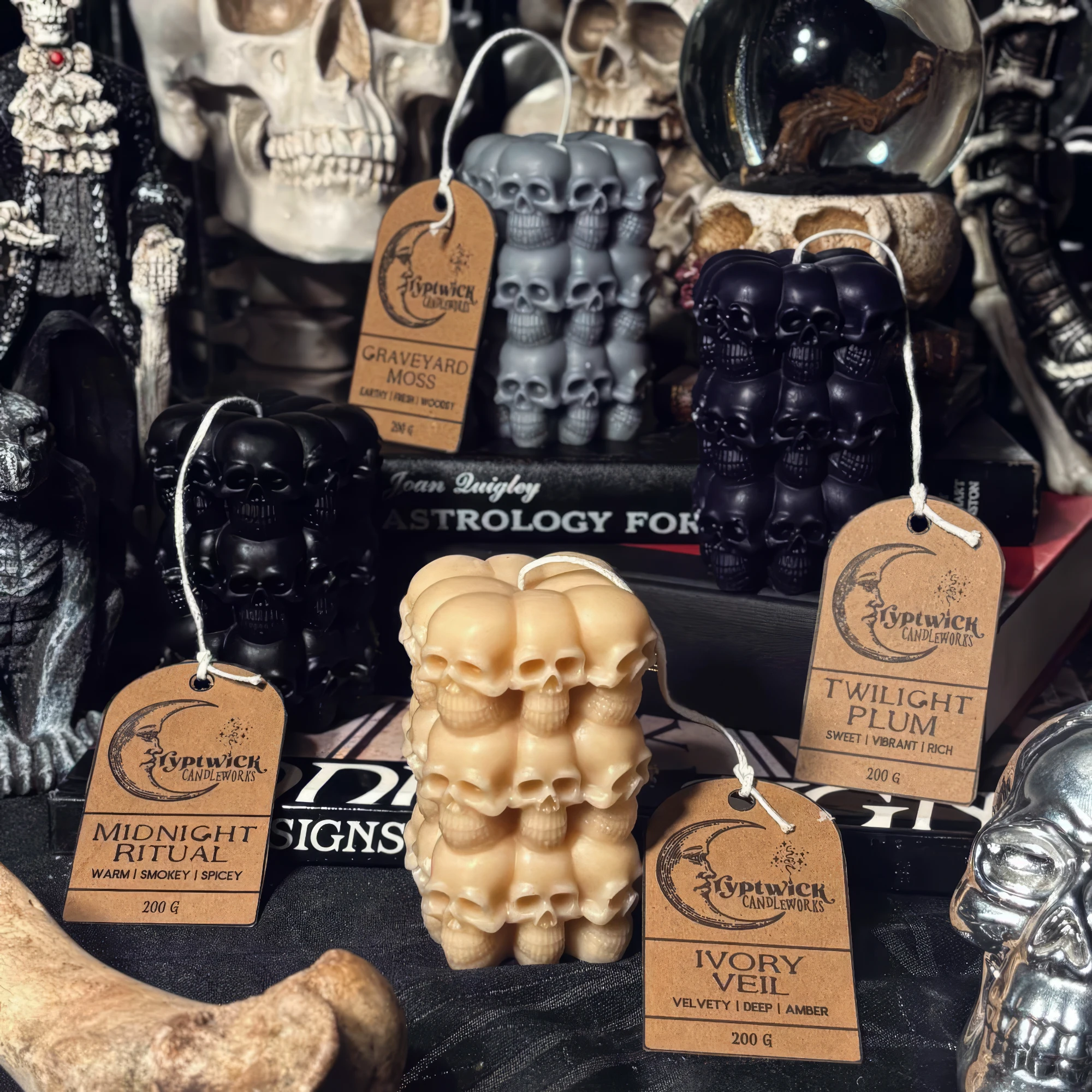 Grim totem candles — stacked-skull pillars in ivory, black, grey and deep plum — each tagged with its scent and weight, among skulls and antique books.