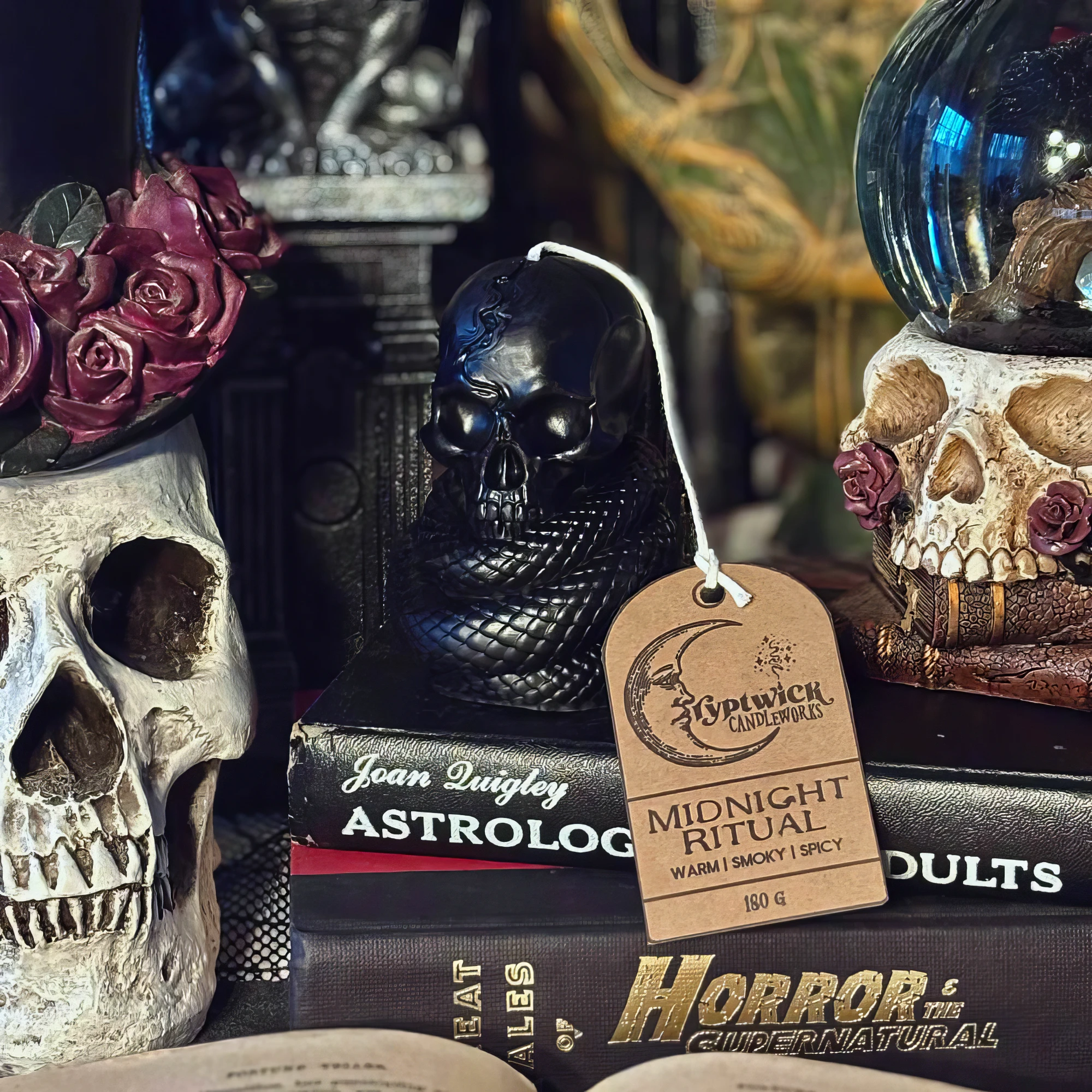 A black serpent-coiled skull candle from the Midnight Ritual scent, tagged in kraft paper, staged among dried roses, a bone skull, antique occult books and a crystal ball.