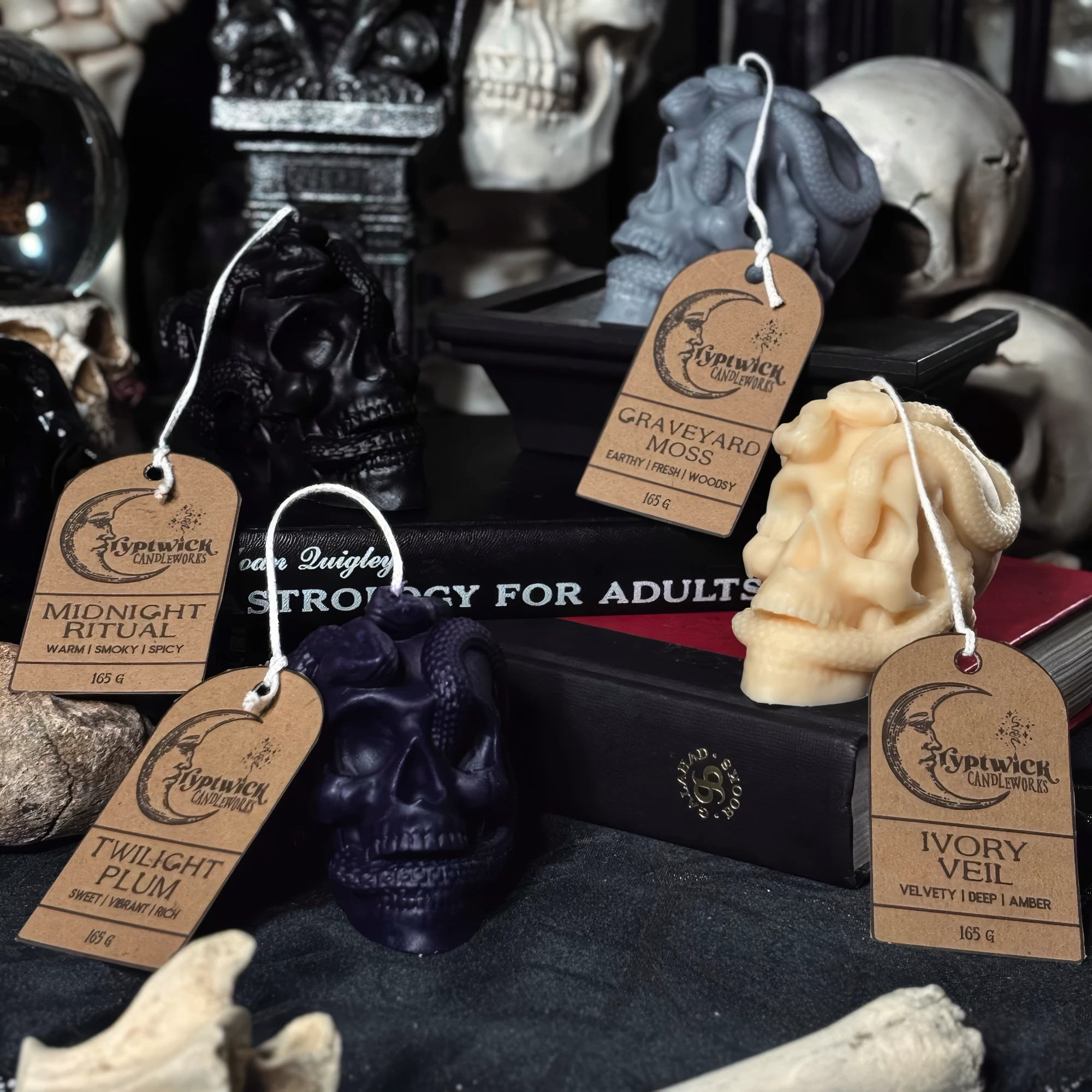 Serpent-coiled skull candles in black, ivory, grey and plum, each with a Cryptwick kraft hang tag, staged among skulls, dried roses and a crystal ball.